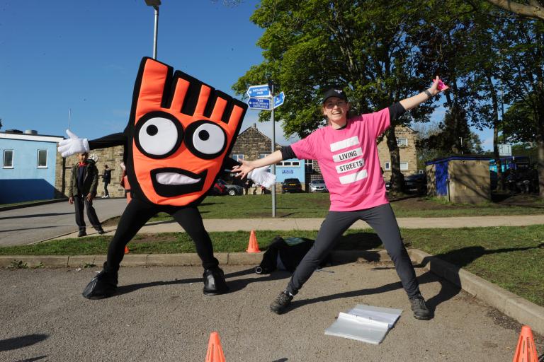 A person is dressed as an orange mascot with large cartoon googly eyes, next to them stands a person in a pink t shirt with the Living Streets logo on it. Both stand with arms and legs wide smiling at the camera