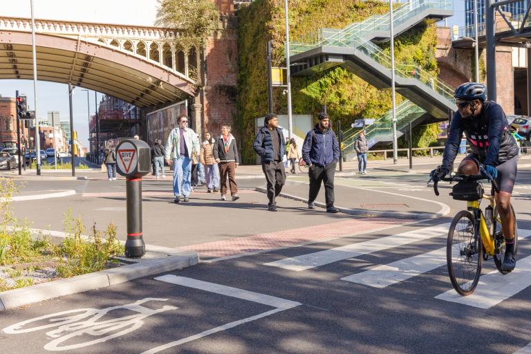 Pedestrians approaching a zebra crossing on a busy urban road with a cyclist is passing