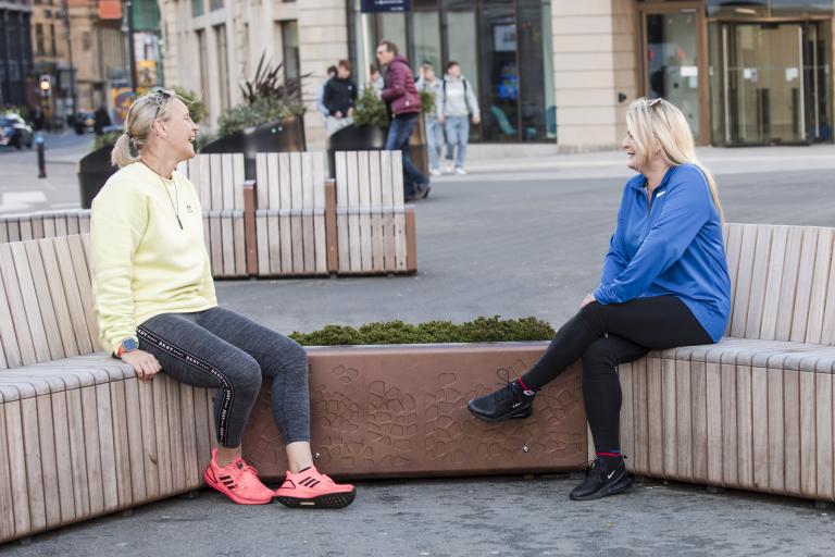 Two women sit on street furniture in a pedestrianised space