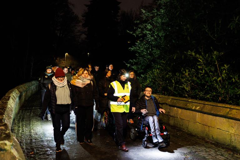 A group of people walking and wheeling along a path in a wooded area after dark. Some are wearing high vis.