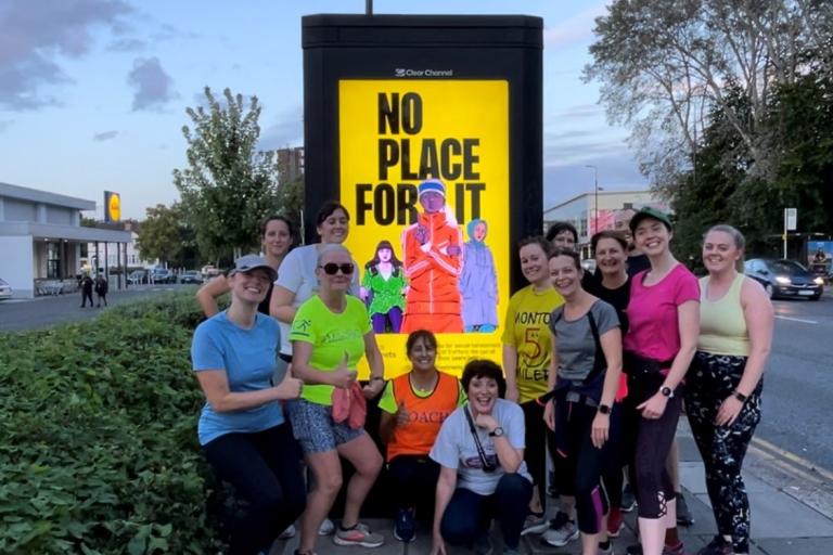 A group of women in active clothing pose next to a billboard on a pavement that reads 'No place for it'.