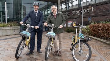 Simon Lightwood and Chris Boardman stood next to e-bikes in front of the entrance to Stockport railway station.