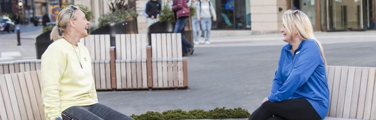 Two women sit on street furniture in a pedestrianised space